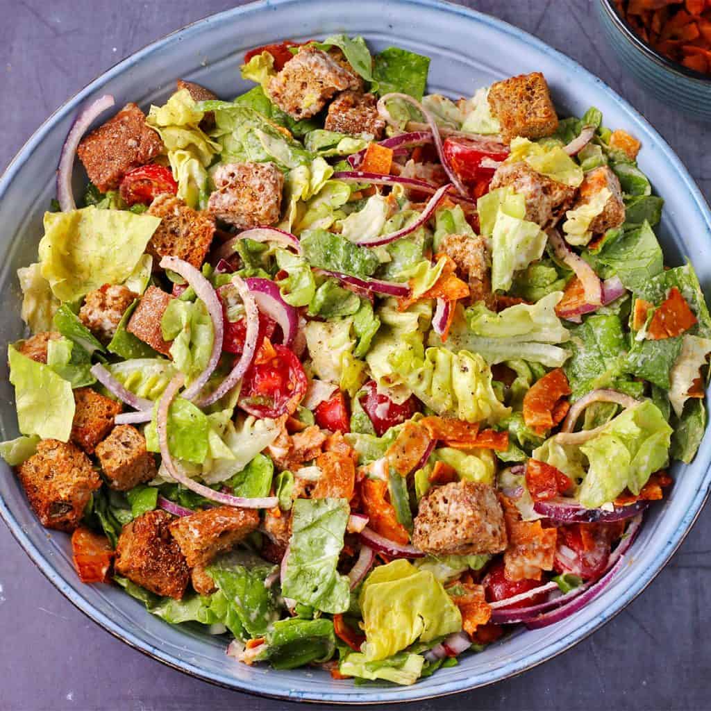 Overhead shot of vegan Caesar salad with chopped lettuce, sliced cherry tomatoes, sliced red onions, coconut bacon, croutons and dressing in blue bowl