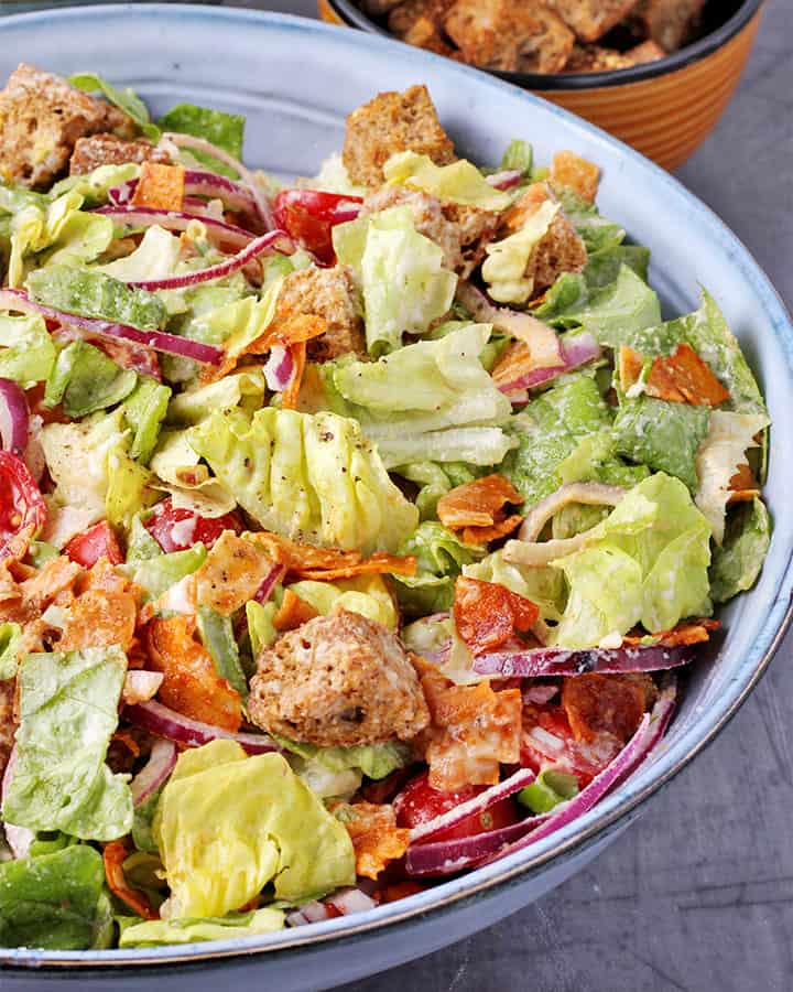 Close up shot of blue bowl with chopped lettuce, sliced cherry tomatoes, sliced red onions, croutons, coconut bacon and vegan Caesar salad dressing
