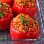Whole red peppers stuffed with barley and white beans and tomato sauce garnished with chopped parsley in glass baking dish