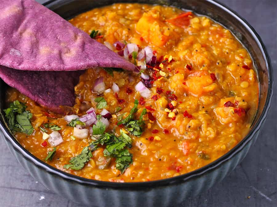 purple flat bread is dipped into post of cooked red lentils and sweet potatoes in blue bowl with chopped cilantro and diced red onions.