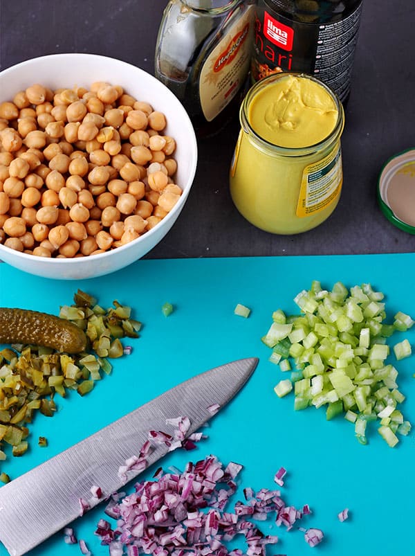dice red onions with knife, diced celery and pickles, bowl of chickpeas and jars of mustard, tamari and maple syrup
