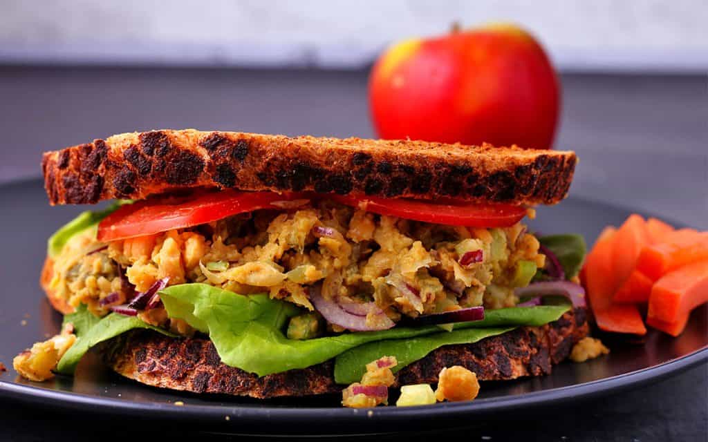 Chickpea salad sandwich with 2 slices of bread, lettuce leaves, red onion slices and tomato slices on black plate with carrot sticks and a red apple