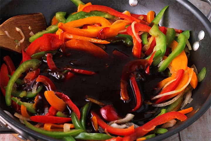 Sweet and sour sauce is added to red, gree, and yellow pepper slices in wok.