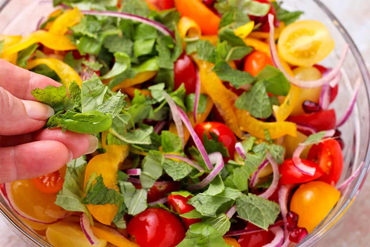 fresh basil and mind is added to salad of tomatoes, red onions, pomegranate seeds, and yellow pepper