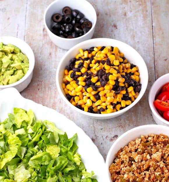 Lettuce, chorizo tofu, black beans and corn, sliced cherry tomatoes, diced avocados, and sliced black olives in white bowls.