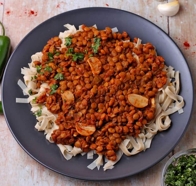 Lentils in Diavolo sauce are served over rice noodles on a black plate.