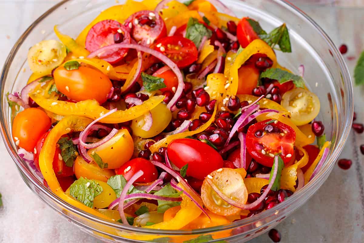 A glass bowl with tomato and pomegranate salad with yellow pepper, red onion, fresh basil, fresh mint, and Za'atar spice blend