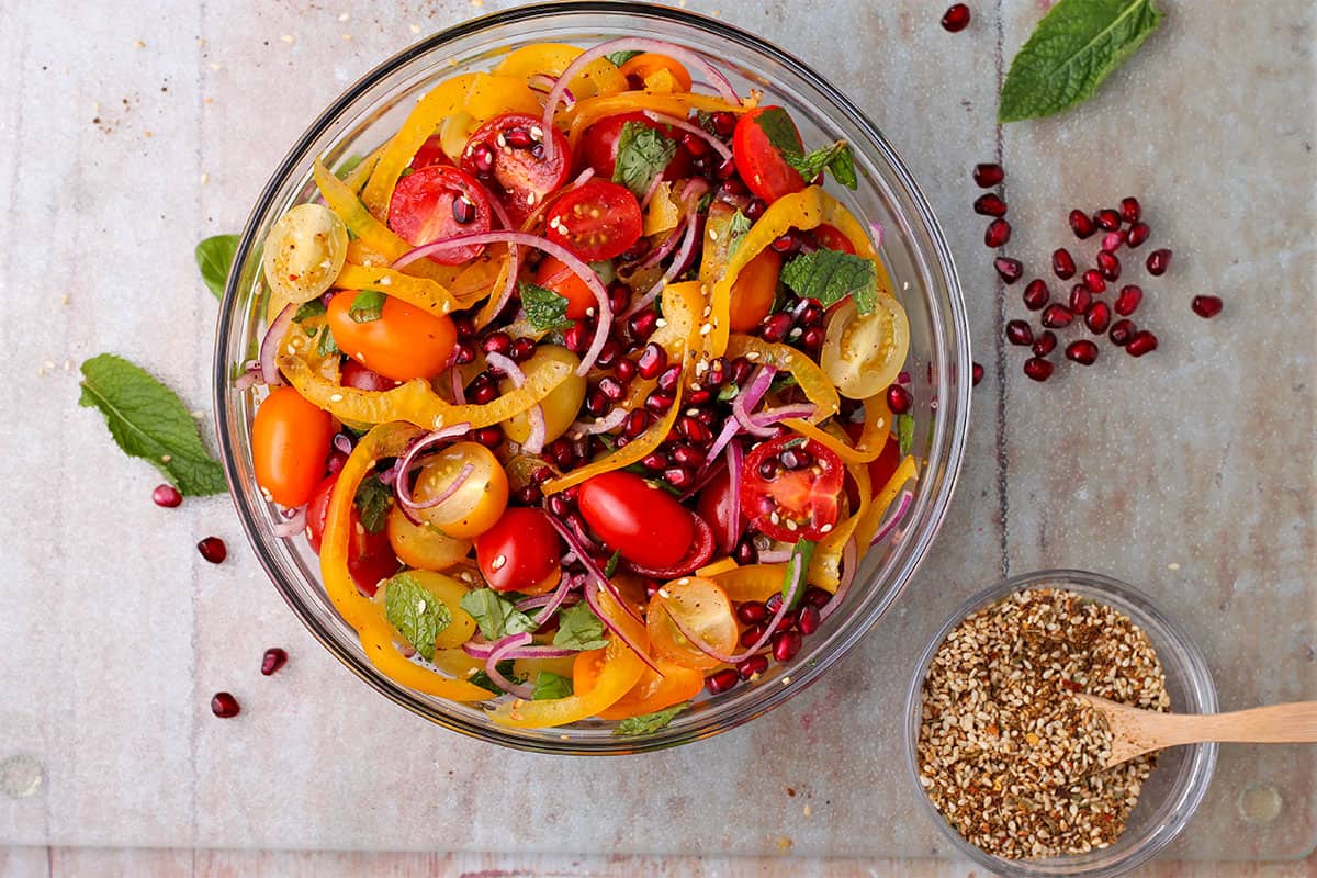 tomato and pomegranate salad in glass bowl with sliced yellow pepper and red onion and fresh basil and mint and bowl of Za'atar spice blend with small wooden spoon.