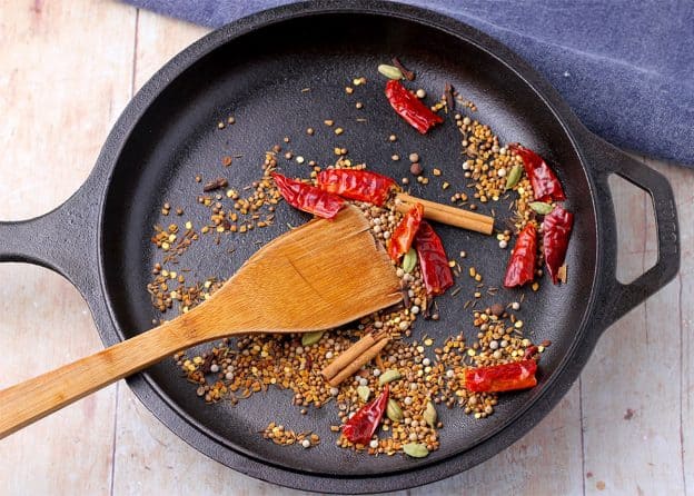 Black cast iron pan filled with dried red chilies, a broken cinnamon stick, allspice berries, cardamom seeds, coriander seeds, cumin seeds, fenugreek seeds, and white peppercorns