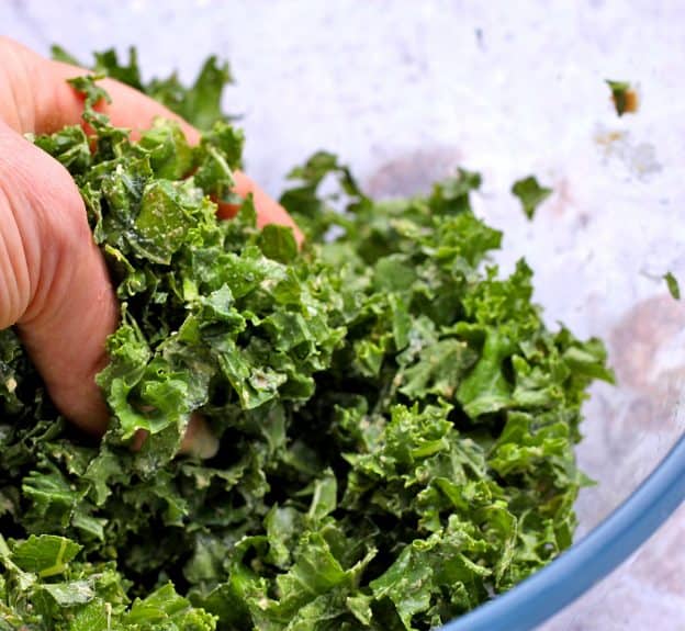 A handful of kale is massaged by hand with Tahini over a glass bowl.