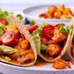 Tacos with roasted cauliflower, Chipotle cream, shredded lettuce, and sliced tomatoes are stacked side-by-side.