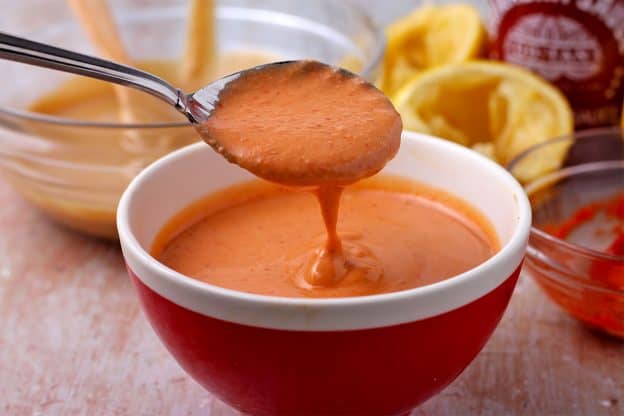 A spoonful of sriracha-tahini dressing is held over a red bowl filled with dressing.