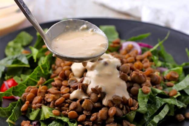 A ladle of lemon tahini salad dressing is poured over a green salad with cooked lentils.