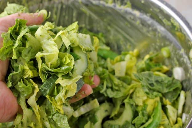 Chopped romaine lettuce, with diced cucumber, scallions, and snap peas is mixed with green goddess dressing with a hand.