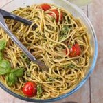 Spaghetti al pesto with tomatoes, fresh basil leaves, and tongs in a glass bowl.