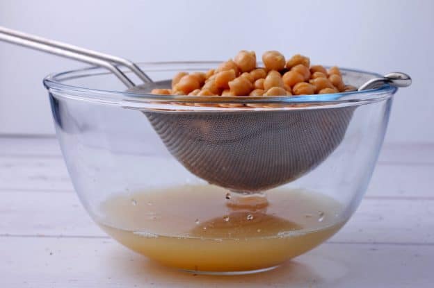 Cooked chickpeas are drained in a strainer over a glass bowl.
