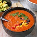 Creamy vegan tortilla soup in a black bowl with corn, black beans, cilantro, and diced tomatoes.