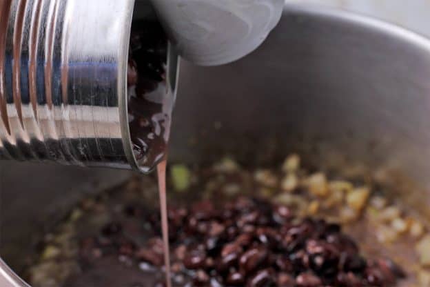 A can of black beans and liquid is poured into a pan with onions and garlic.
