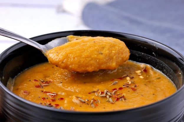 A spoon of carrot and lentil soup with mixed spices is lifted over a soup bowl.