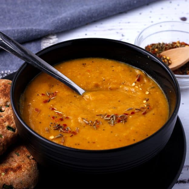 Spiced carrot and lentil soup with toasted cumin seeds, chili flakes and mustard seeds in a black bowl.