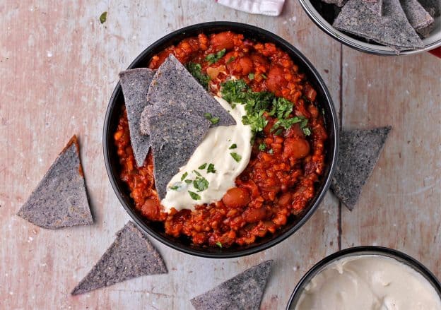 Plant-based chili in a black bowl with a bowl of vegan sour cream and blue corn tortilla chips.