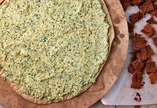 A pizza crust with artichoke pesto spread and plant-based bacon on the side.
