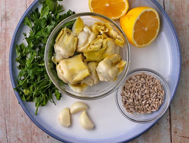 A blue plate with peeled garlic cloves, a small bowl of sunflower seeds, fresh parsley, cut lemon halves, and artichoke hearts.