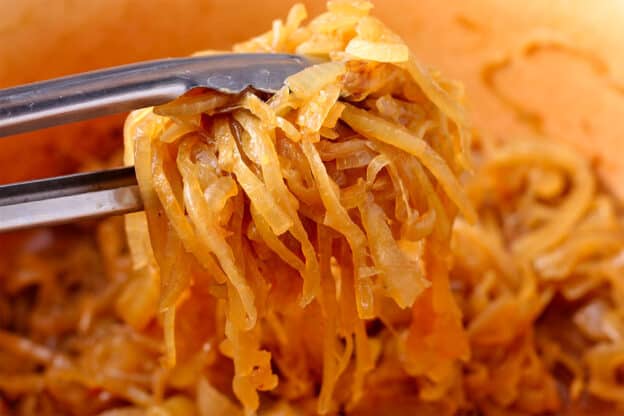 Caramelized onions are held with tongs over a pot of onions.