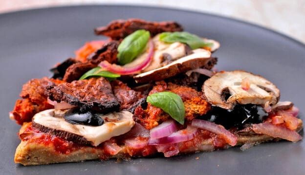 A slice of quinoa crust pizza with mushrooms and plant-based bacon on a gray plate.