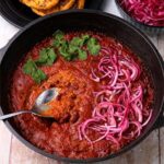 rajma masala, kidney bean curry in a black cast iron pot with a spoon is topped with pickled red onions and cilantro.