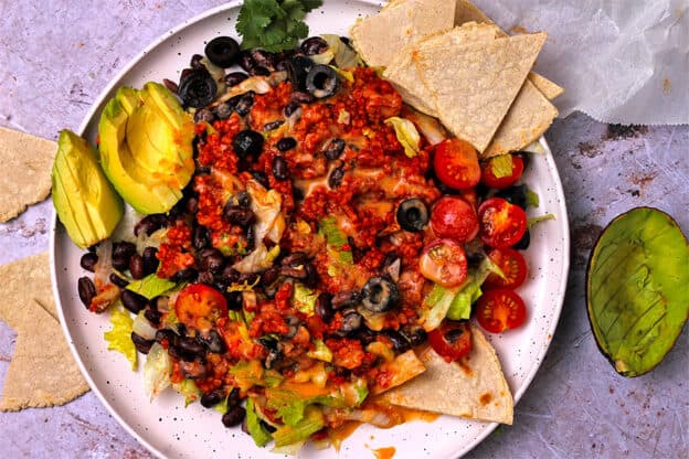 A taco salad on a white plate with chipotle dressing, bulgur taco meat, black olives, lettuce, tomatoes, avocados, tortilla chips, and dressing.