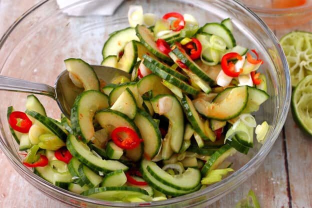 A salad with sliced cucumbers, sliced red chili, scallions, and a chili paste and lime dressing is mixed with a spoon.