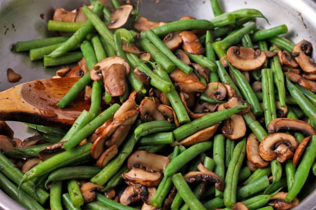 Green beans and mushrooms are cooked in a skillet with a wooden spoon.