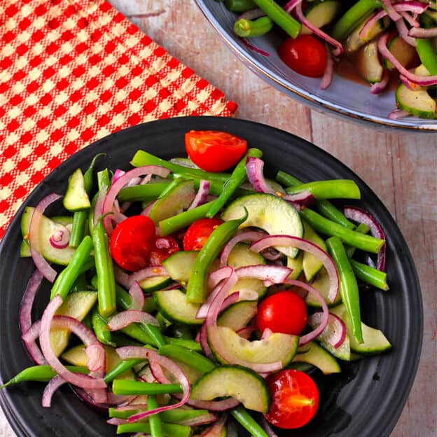 A salad with green beans, cucumbers, cherry tomatoes, and red onions on a black plate.