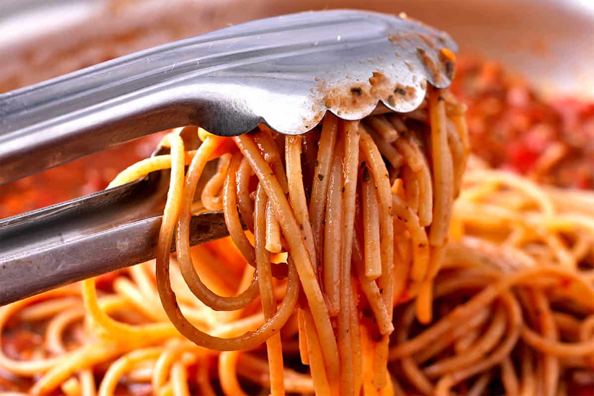 Cooked spaghetti mixed with arrabbiata sauce using tongs.