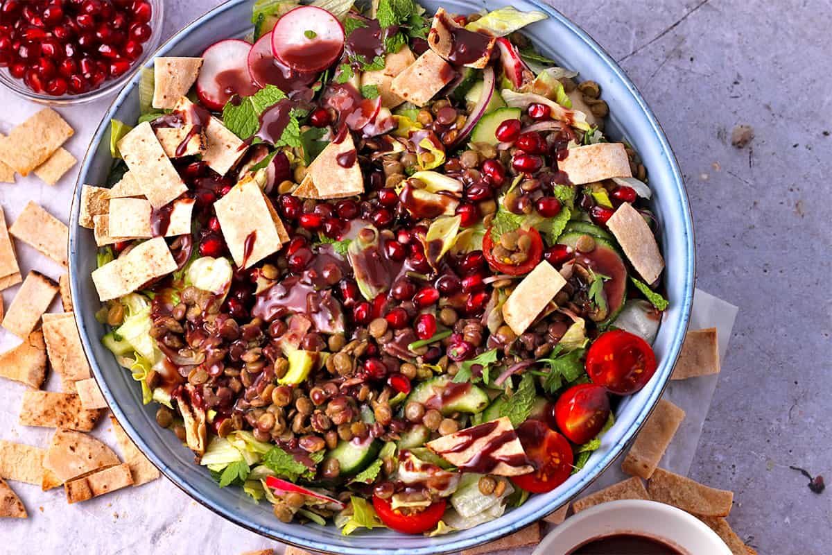 A salad of cooked lentils, pomegranate, tomatoes, pita chips, red onions, lettuce, and dressing.