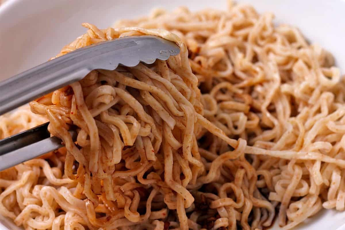 Noodles are mixed with dressing using tongs.