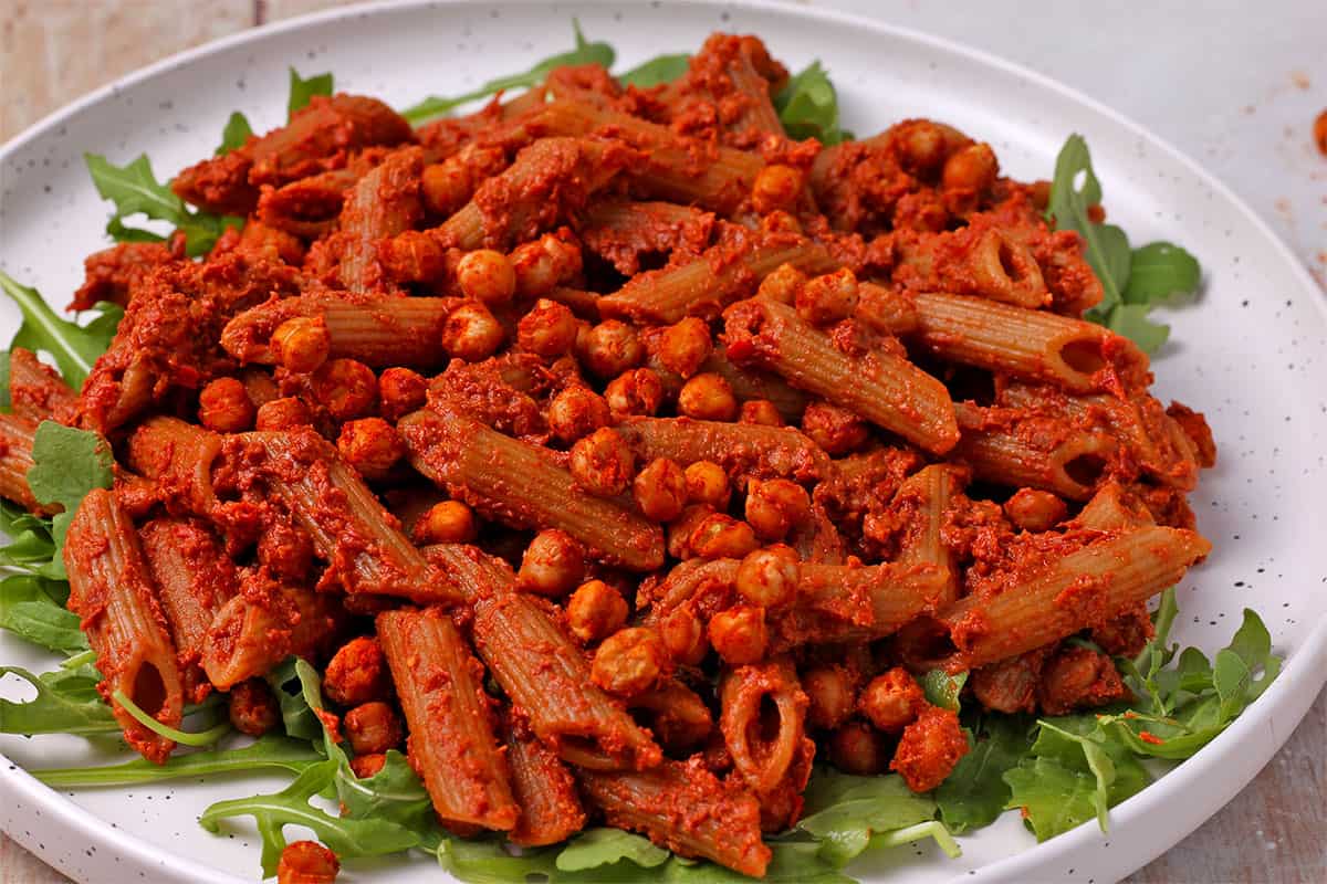 A bed of arugula with pasta mixed with red pesto and topped with seasoned chickpeas.