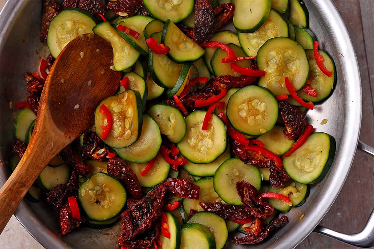 Zucchini, sundried tomatoes, and red chili are sautéed in a pan.