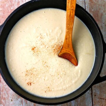 Vegan bechamel, creamy white sauce is presented in a black pan with a wooden spoon.