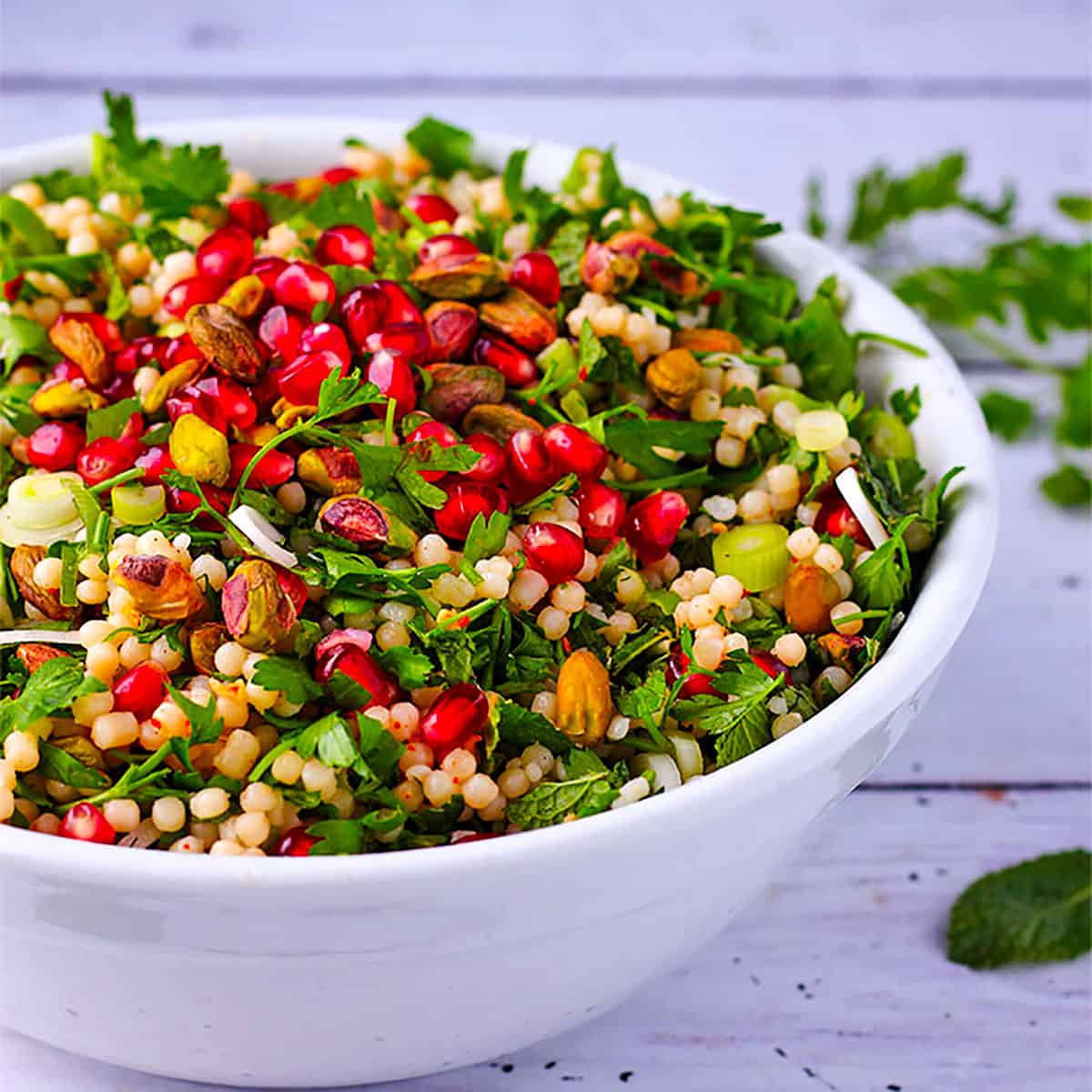 A white bowl filled with cooked Israeli couscous, pomegranate, herbs, scallions, and pistachios mixed in a white bowl.